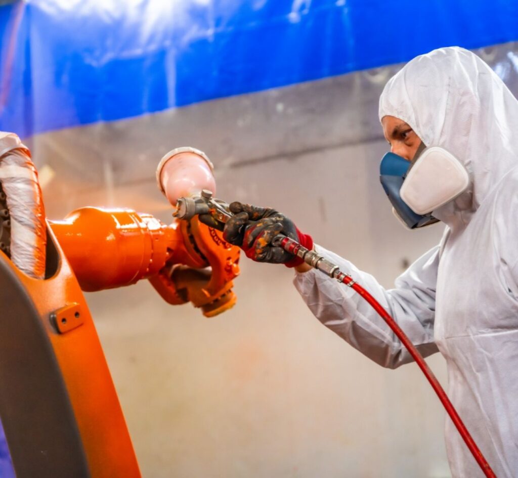 Medewerker in beschermende kleding brengt een oranje coating aan op een industrieel metalen onderdeel met een spuitpistool