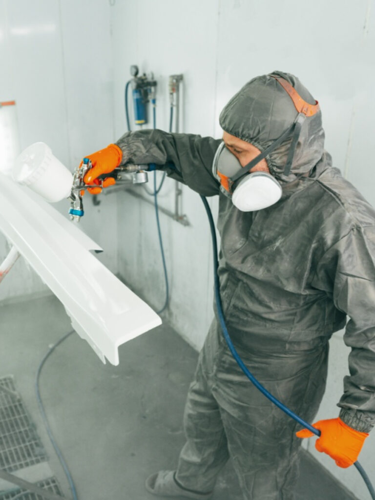 Medewerker in beschermende kleding spuit een witte coating op een metalen profiel in een professionele spuitcabine