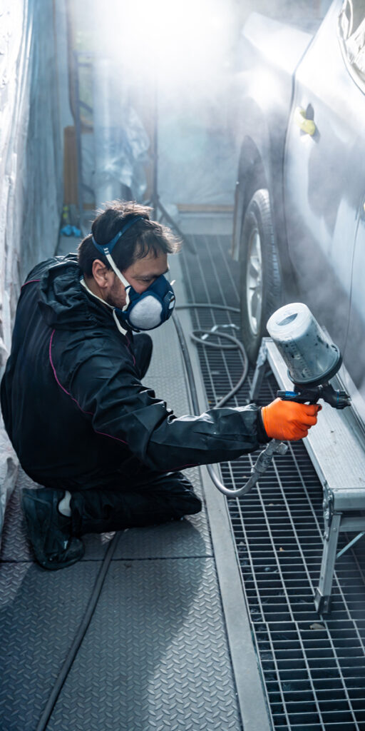 Medewerker met spuitpistool en beschermingsmasker brengt coating aan op een auto in een professionele spuitcabine