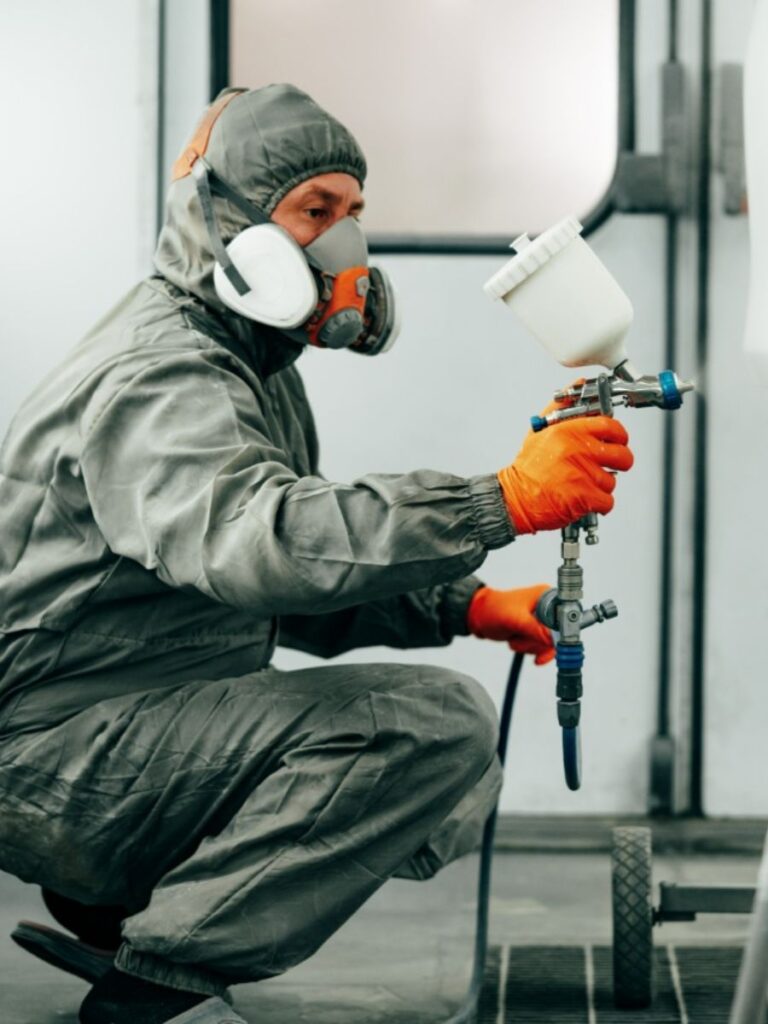Medewerker in beschermende kleding en ademmasker bezig met professioneel spuitcoaten in een industriële spuitcabine
